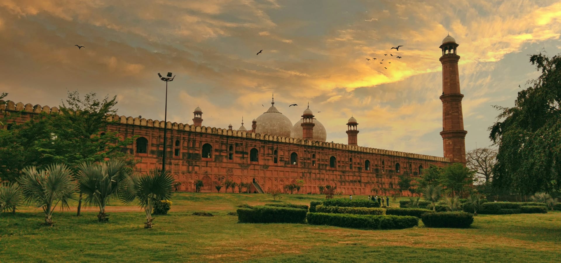 The exterior of the Badshahi Mosque in Lahore, featuring red sandstone walls, white marble domes, and tall minarets under a dramatic sky.