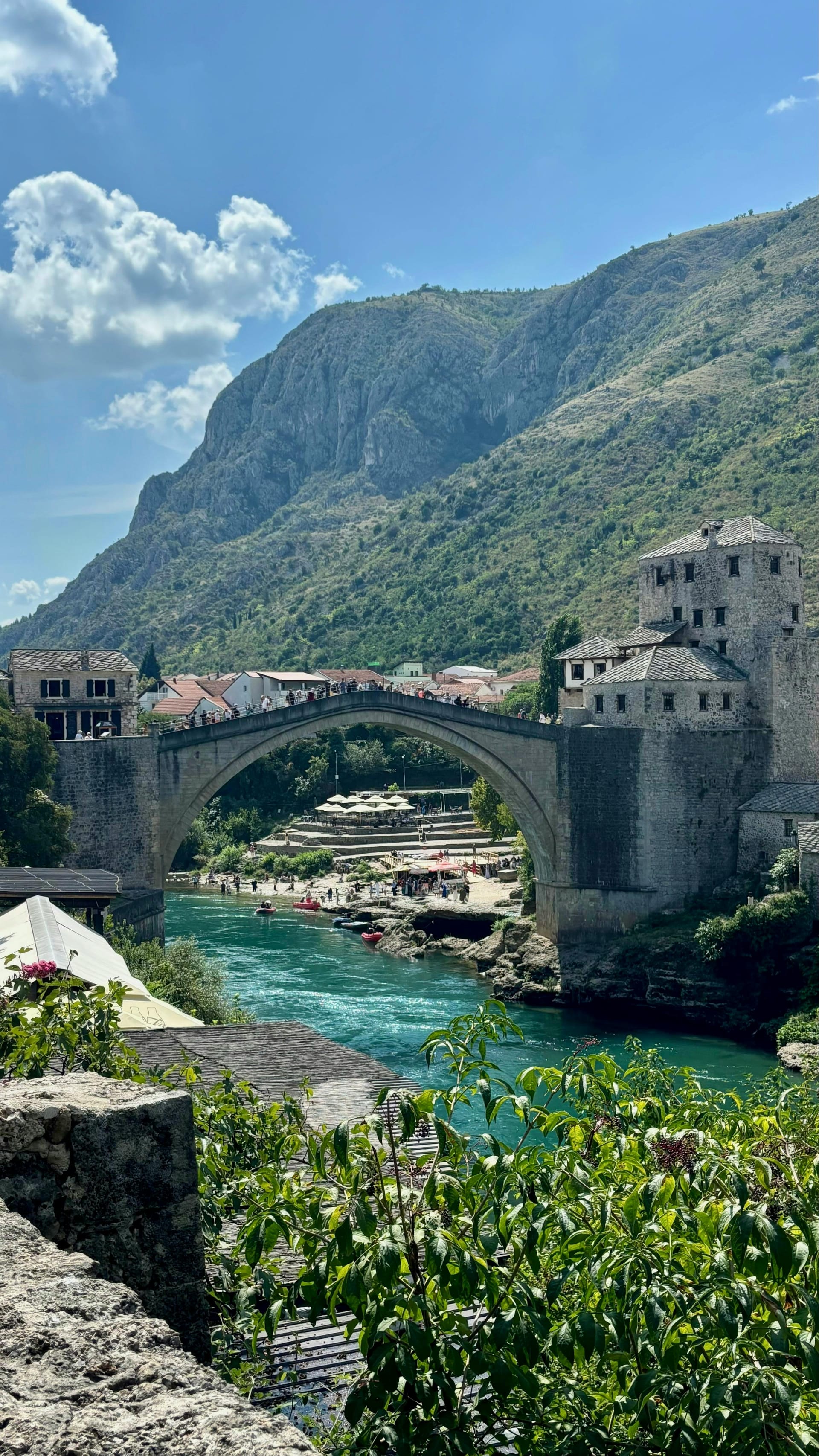 The historic Stari Most stone bridge spanning the green Neretva River in Mostar, with stone buildings on the banks.