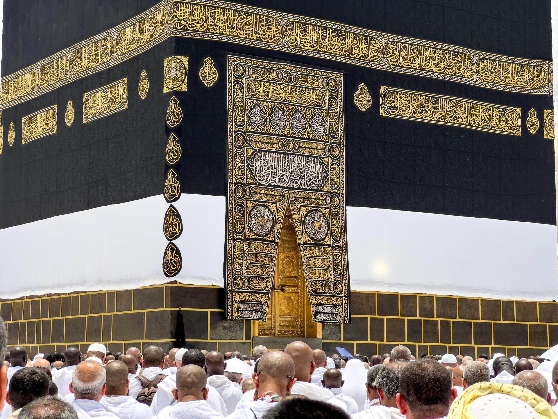 A close-up view of the gold Quranic calligraphy on the black Kiswah cloth of the Kaaba, with pilgrims standing below.