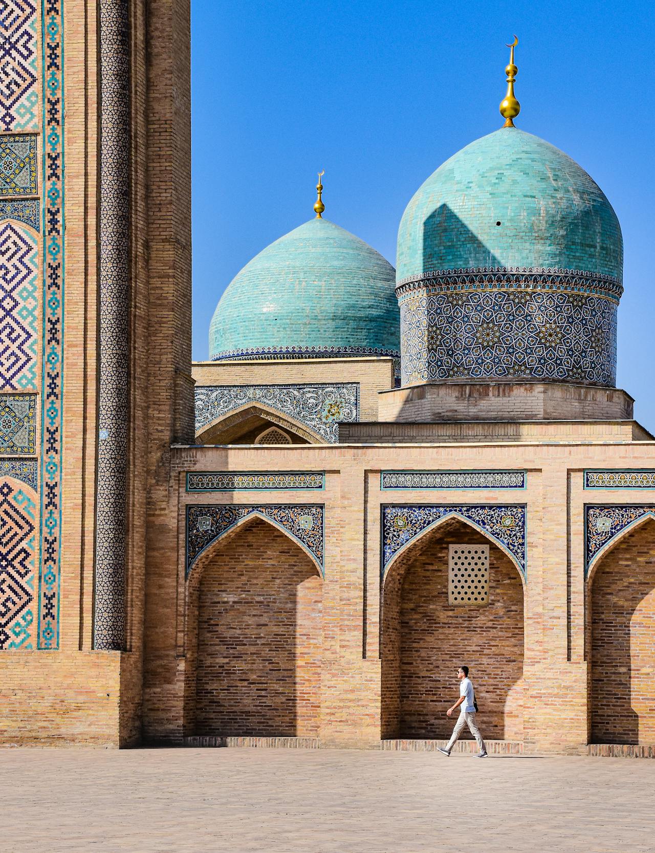 person walking past the intricate blue and turquoise geometric tile work on the tall facade of a historic building in Samarkand.
